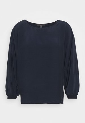 Blouse bleu marin en tissu lisse, avec un col rond et des manches longues bouffantes dotées de poignets ajustés. Coupe droite à l'ourlet.