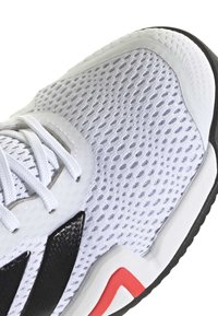 White mesh athletic shoe with black and red accents. Features include a textured surface, reinforced toe, and lacing over the tongue.