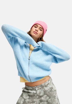 Junge Person mit rosa Mütze, hellblauer Zip-Jacke, geblümter grauer Hose und erhobenen Armen vor einfarbigem Hintergrund.