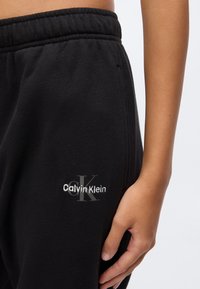 Κοντινή λήψη της μέσης και του χεριού ενός ατόμου που φοράει μαύρο παντελόνι φόρμας Calvin Klein με ελαστική μέση και λογότυπο στο αριστερό ισχίο.