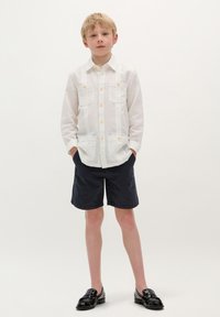 Witte blouse met knopen en twee borstzakken, gecombineerd met zwarte shorts en glanzende zwarte loafers. Eenvoudig, casual ontwerp met een lichte textuur.
