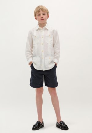 Camicia bianca con bottoni, dotata di due tasche sul petto, abbinata a pantaloni corti neri e mocassini neri lucidi. Design semplice e casual con una leggerissima trama.