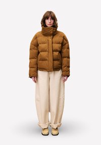 Persoon die tegen een witte achtergrond staat en een bruine pufferjack, brede beige broeken met omgeslagen enkels en beige schoenen draagt.