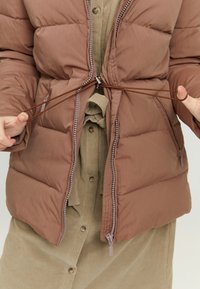 Brown puffer jacket with a zipped front, adjustable drawstring at the waist, and a textured olive green shirt underneath.