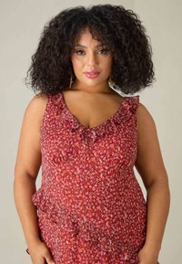 Rödblommig klänning med volang vid halslinning och nederkant, som har en figurnära passform och mjuk tyg. Modellen har axellångt lockigt hår.