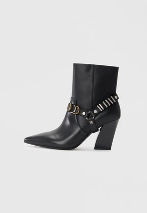 Black leather ankle boot with pointed toe, stacked heel, and decorative strap featuring metal hardware and studs. Smooth texture.