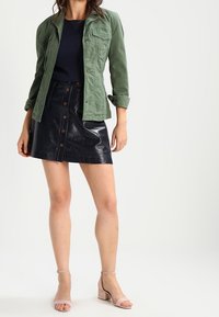 Grön bomullskavaj över en marinblå topp, kombinerat med en svart fusk läderkjol med frambutton, och ljusrosa blockklack sandaler.