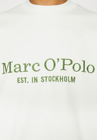 T-shirt en coton blanc avec texte brodé vert : "Marc O'Polo EST. À STOCKHOLM." Design classique à col rond.