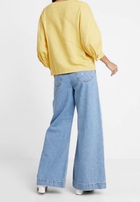 Geel, losvallend blouse met opgeblazen mouwen en knoopaccenten, gecombineerd met lichtblauwe, wijd uitlopende jeans met achterzakken.