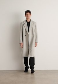 Palton gri deschis din lână cu guler tip șal, design cu două rânduri de nasturi, mâneci lungi și deschideri laterale, asortat cu cămașă și pantaloni negri.