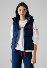 Ναυτικό μπλε γιλέκο με κουκούλα, διαθέτοντας ραφές και κόκκινο φερμουάρ, φορεμένο πάνω από ένα λευκό φούτερ με γραφικό σχέδιο.