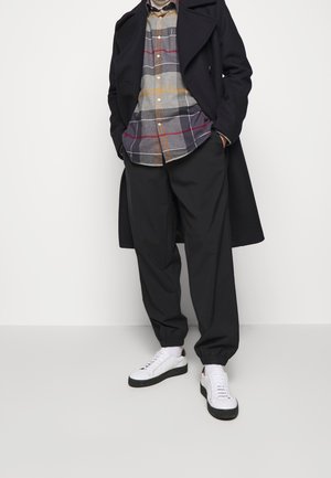 Zwarte wollen jas over een grijs geruit overhemden met oranje en rode accenten. Losse zwarte broek en witte sneakers met zwarte zolen.