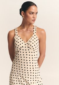 Crème kleurige jurk met zwarte stippen. Heeft ruches op de schouderbanden, een V-halslijn en een getailleerd silhouet met een zachte textuur.