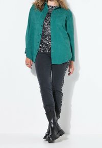 Chemise en daim teal, haut graphique noir, jeans foncés avec des clous en argent et bottes noires chunky. Textures lisses, fermeture à bouton sur la chemise.