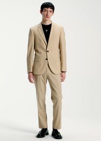 Beež komplekt, millel on ühe rea nööbid, notsitud kraega jakk, kaks taskut ja sobivad püksid. Must ümmarguse kaelusega särk all, kantud mustade kingadega.