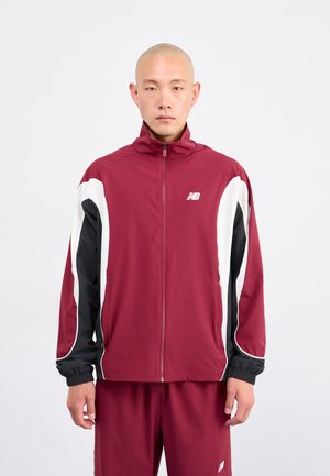 Maronfarbige Trainingsjacke mit schwarz-weißen Akzenten, hohem Kragen, Reißverschluss vorne, elastischen Bündchen und passenden maronfarbenen Hosen. Der Stoff wirkt glatt und leicht.