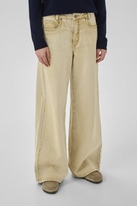 Person, der beigefarbene Jeans mit weitem Bein, braune Mokassin-Schuhe und ein marineblaues Langarmshirt trägt, steht vor einem schlichten grauen Hintergrund.