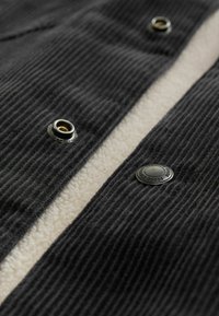 Tela de pana negra con un forro de felpa beig, que presenta botones de presión metálicos y una textura acanalada. Enfocándose en una construcción durable y contraste de colores.