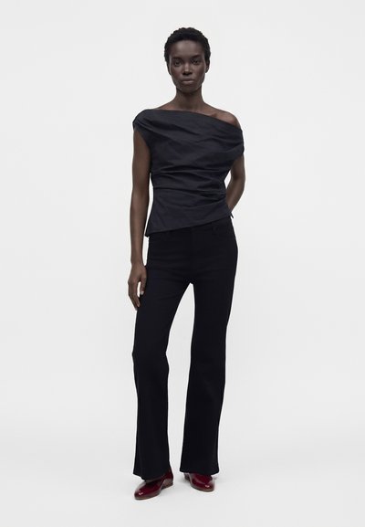 Femme debout portant un haut noir épaules dénudées, un pantalon noir évasé et des chaussures rouge foncé sur un fond clair uni.