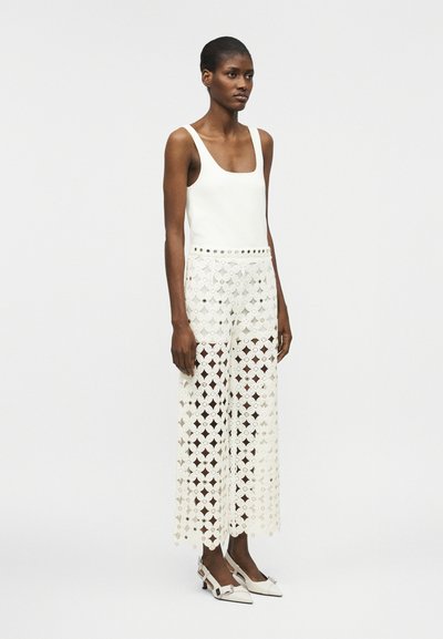 Femme portant un haut blanc sans manches associé à un pantalon large en dentelle blanche taille haute et des escarpins slingback pointus assortis.