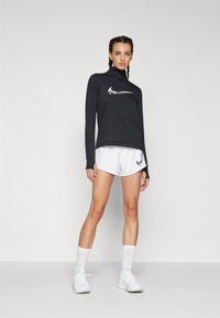 Black long-sleeve top with a high collar and white logo, paired with white shorts featuring a black logo. White sneakers and socks complete the look.