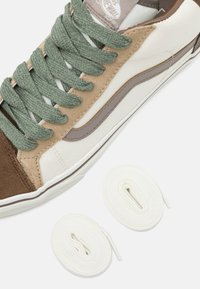 Zapatilla beige, marrón y blanca con cordones verdes, junto a dos cordones blancos bien enrollados, sobre un fondo blanco.