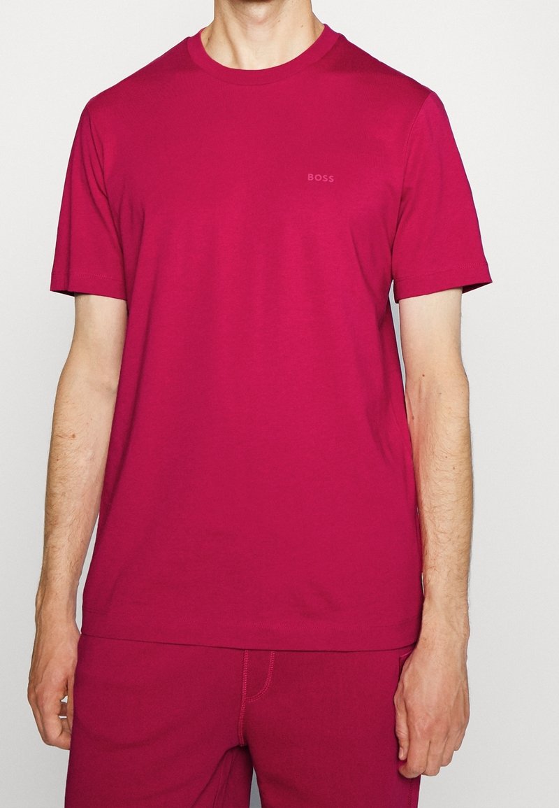 Homme portant un T-shirt à manches courtes magenta uni et un pantalon assorti avec un logo subtil "BOSS" sur la poitrine gauche, debout devant un fond blanc.