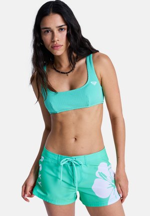 Młoda kobieta o długich ciemnych włosach, ubrana w turkusowy teksturowany top bikini oraz pasujące krótkie spodenki kąpielowe z białym nadrukiem kwiatów hibiskusa.