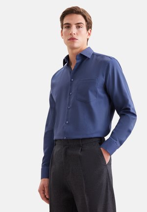 Jeune homme portant une chemise bleue à boutons avec une poche poitrine et un pantalon gris foncé, debout avec une main dans sa poche.