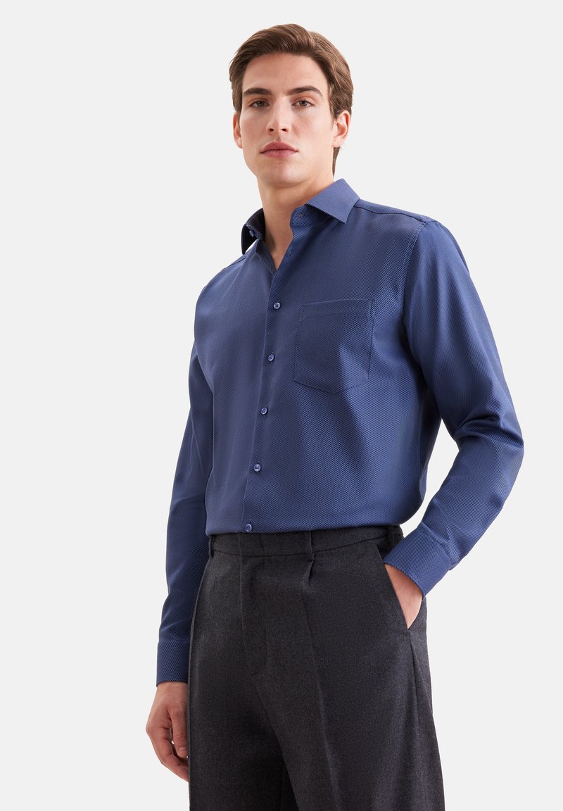 Jeune homme portant une chemise bleue à boutons avec une poche poitrine et un pantalon gris foncé, debout avec une main dans sa poche.