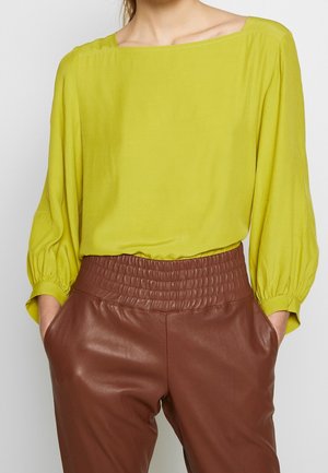 Vrouw draagt een limoengroene blouse met lange mouwen, ingestopt in een hooggetailleerde bruine leren broek, handen in de zakken.