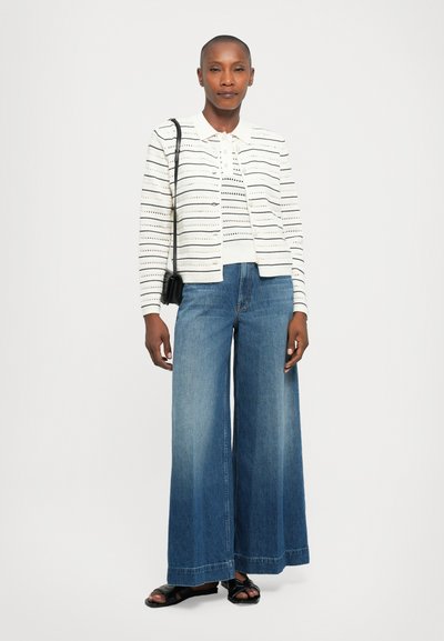 Femme portant un cardigan rayé blanc et une chemise polo, un jean large bleu, un sac bandoulière noir et des sandales noires, se tenant devant un fond blanc.