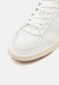 Balti oda sportiniai bateliai su perforuota pirštų sritimi, plokščiais baltais raišteliais, tekstūruotais akcentais ir šviesios spalvos gumele.