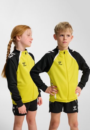 Młodzieżowe zestawy sportowe z jasnżółtą kurtką zapinaną na zamek z czarnymi akcentami oraz szortami, wykonane z gładkiego, elastycznego materiału.