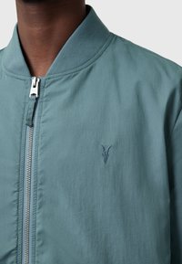 Giacca bomber di un verde acqua chiaro con una texture liscia, dotata di una cerniera frontale argento e un piccolo logo ricamato sul lato sinistro del petto.