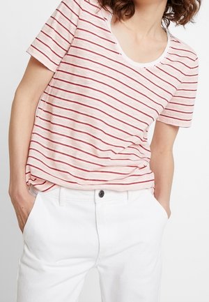 Vrouw met een roze-wit horizontaal gestreept shirt met korte mouwen, ingestopt in witte broek, handen in de zakken, met krullend haar.