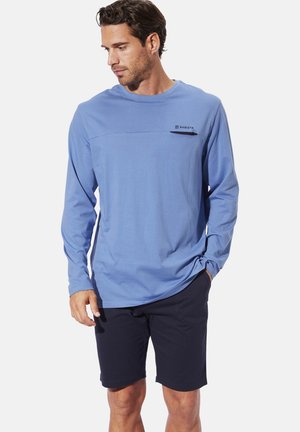 Langärmliges, hellblaues Hemd aus glattem Stoff, mit einer horizontalen Naht und einem kleinen schwarzen Logo. Kombiniert mit dunklen Shorts.