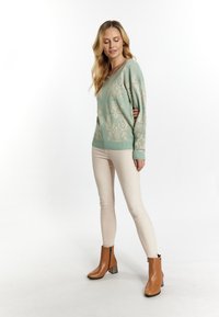 Hellgrüner Pullover mit einem strukturierten cremefarbenen Muster, ausgestattet mit einem V-Ausschnitt und gerippten Bündchen, kombiniert mit beigen Slim-Fit-Hosen und braunen Stiefeletten.