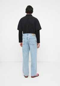 Camisa preta de manga curta, calças jeans largas em azul claro com bolsos traseiros, e sandálias com listras vermelhas, vistas de trás contra um fundo simples.
