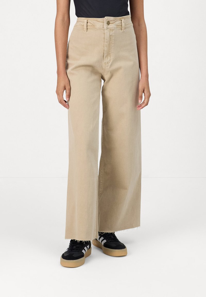 Beige Jeans im weiten Schnitt mit hoher Taille und fransigem Saum, ausgestattet mit einer vorderen Knopfverschluss und Seitentaschen. Getragen mit schwarzen Sneakers.