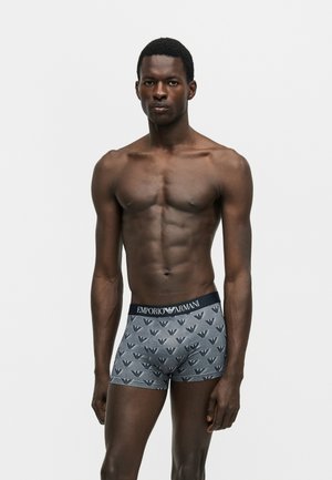 Atletyczny męski model stojący, ubrany w szare bokserki Emporio Armani z wzorem logo na gładkim jasnym tle.