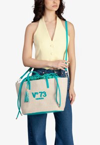 Borsa a rete beige con dettagli turchesi, comprese le maniglie e il logo. Presenta una nappina e una chiusura con coulisse nella parte superiore.