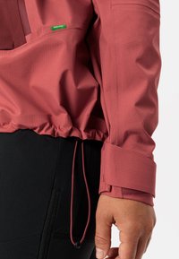 Giacca rossa di peso medio con orlo regolabile, polsini con bottone e tessuto testurizzato. Etichetta verde sul lato sinistro superiore. Indossata sopra abbigliamento nero.
