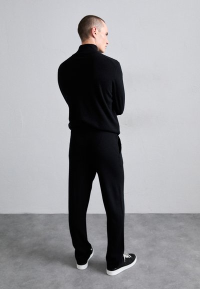 Black knit polo and matching pants, featuring a relaxed fit. Model stands with back to the camera, wearing black sneakers on a gray floor.
