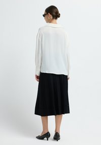 Blouse blanche à manches longues avec un col, assortie à une jupe plissée noire. Des chaussures à talons noires complètent la tenue.