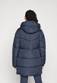 Marineblauwe gewatteerde jas met een capuchon, met doorgestikte secties en een iets langere achterkant. Het materiaal lijkt glad en geïsoleerd.