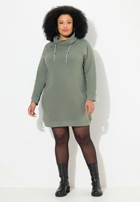 Vestido de algodón verde con escote drapeado, mangas largas y detalles con cordones. Combinado con botas negras hasta la rodilla y medias negras transparentes.