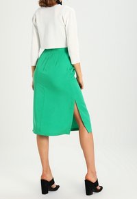 Zelená sukně po kolena s bočním rozparkem, vyrobená z hladké látky. Kombinovaná s přiléhavým bílým krátkým topem a černými sandály na vysokém podpatku.