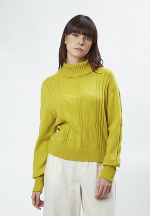 Pull en maille jaune avec un col montant, présentant des motifs en torsades et des poignets côtelés. Porté sur un pantalon clair et ample.