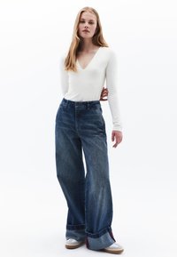 Witte lange mouwtop met een V-hals en blauwe wijde jeans, met een hoge taille en opgerolde manchetten. Model draagt beige sneakers.